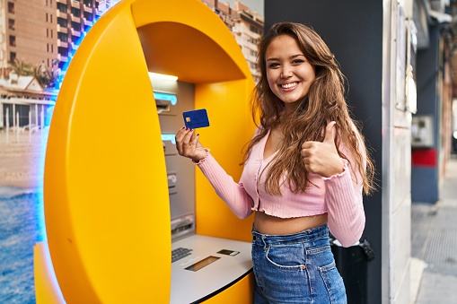 Young hispanic woman taking cash from atm with credit card smiling happy and positive, thumb up doing excellent and approval sign