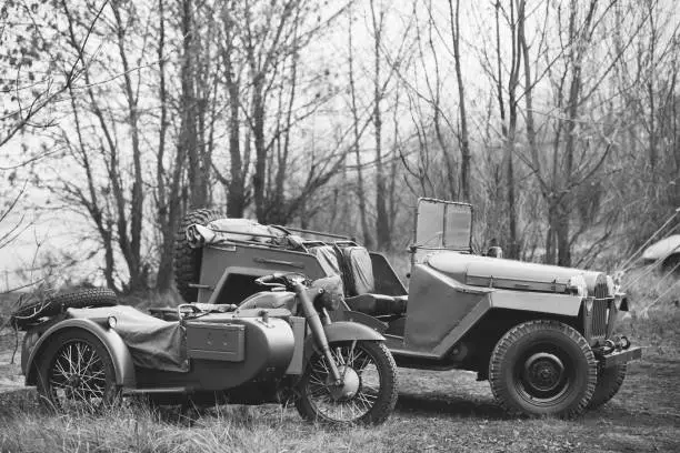 Photo of Old Tricar, Three-Wheeled Gray Motorcycle With A Sidecar Of German Forces and Willys Mb jeep, U.s. Army Truck, 4x4 Parked At Forest During Reconstruction Of Some Fight World War II. Black and white