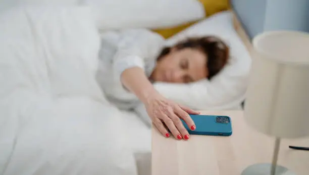 Middle age hispanic woman turning off smartphone alarm lying on bed at bedroom