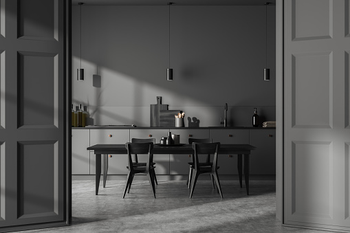 Dark kitchen interior with dining table and chairs, dresser with kitchenware on grey concrete floor. Opened doors leading into cooking area with modern furniture. 3D rendering