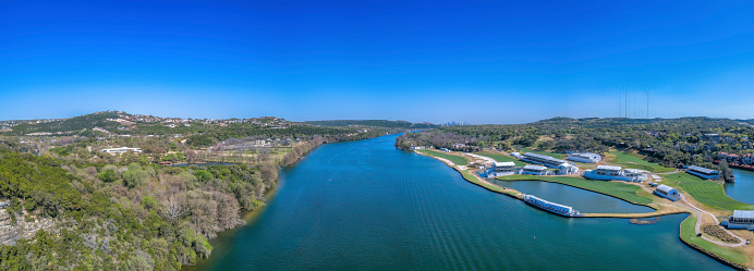 Austin, Texas- Panoramic view of Colorado River against the blue sky background. There are green land with mountain slopes and buildings on the side of the river.