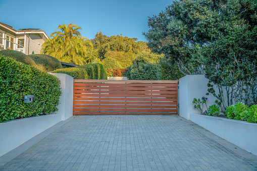 La Jolla, California- Entrance of a private residence with concrete driveway and large wooden gate. There are plants and trees surrounding the driveway and the building on the left.