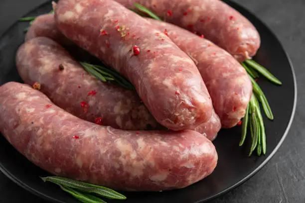 Photo of Raw sausages or bratwurst with spices and rosemary in a plate on black background. Close up