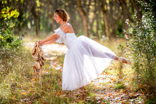 Mature ballet dancer in the forest posing with her leg up and old dance shoes in his hands