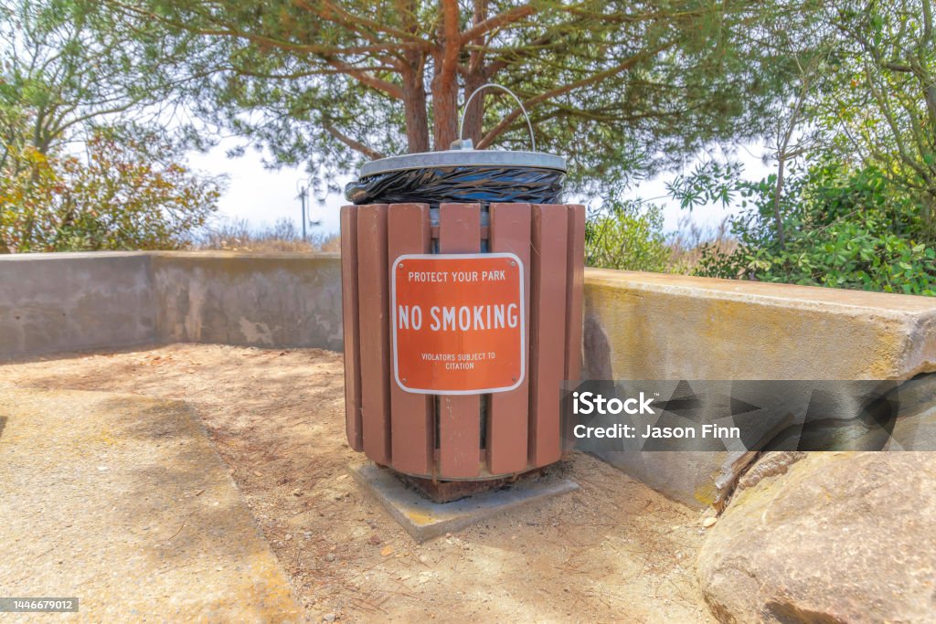 Public trash container with cover and no smoking sign at Laguna Niguel in California Public trash container with cover and no smoking sign at Laguna Niguel in California. Trash container near the concrete wall and large rock against the trees at the back. Architecture Stock Photo