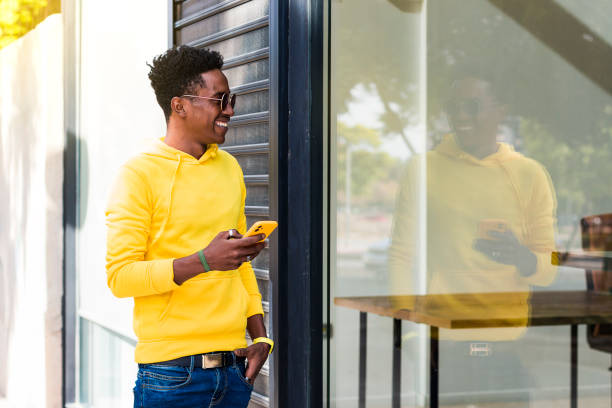 Portrait of a smiling young African man wearing a yellow sweatshirt, jeans and sunglasses looking at the shop window in which he is reflected while holding a cell phone.