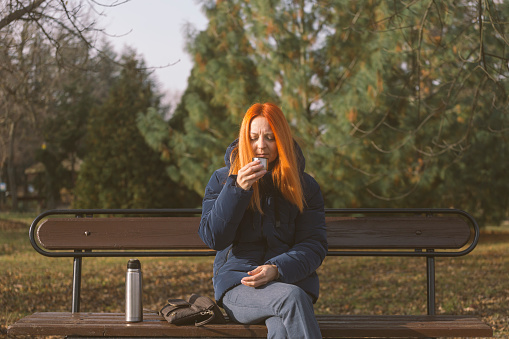 Woman drinking hot tea to warm her outdoors in public park on cold autumn day