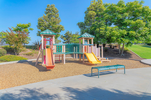 Small playground in a park at San Diego, California. There is a bench on the concrete ground at the front and a view of plants, trees and lawn at the back.