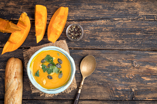 Butternut squash soup in a bowl with pumpkin seeds and fresh butternut squash on wooden background, top view