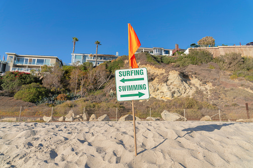 Directional signage near the buildings at San Clemente, Orange County, California. Swimming and surfing signage with orange flag on top against athe view of a slope with buildings on top.