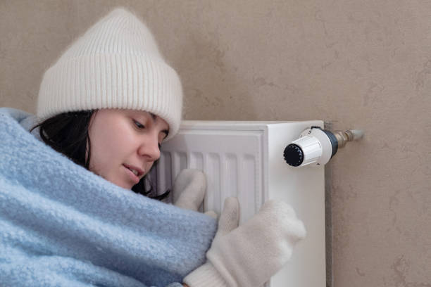 a warmly dressed woman leaned against the radiator and tried to warm herself. increase in the cost of heat stock photo