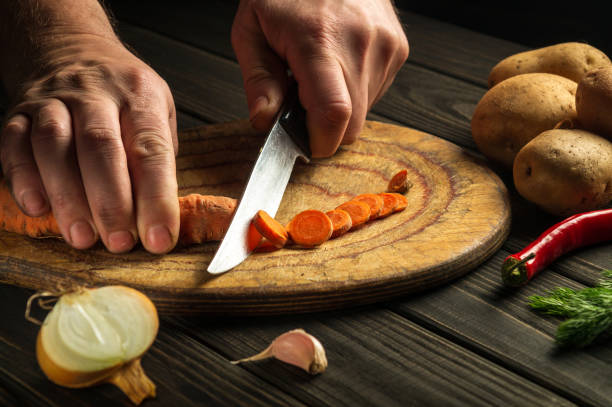 istock Slicing carrots on a wooden board for cooking. Chef hands with a knife while working on the kitchen table. Peasant foods. 1446696229