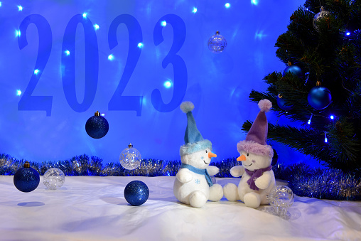 Christmas greeting card with a pair of toy snowmen under Christmas tree and numbers 2023.