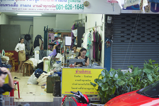 Thai women are sewing in open shop in street Chokchai 4 in Bangkok Ladprao. In background one woman is checking some sewed fashion.  View over street and sidewalk from opposite side into open work place and shop