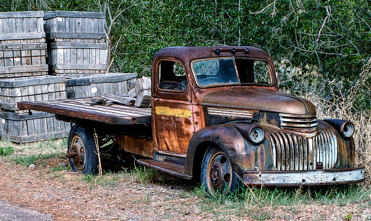 An antique and rusty flat bed truck is seen on the side of a rural road. The truck is missing a windshield and has flat tires but it was once used to transport goods and haul families.