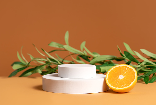 Template for cosmetic product presentation made with white round podium, orange slice and olive tree branch on brown background. Trendy fashion showcase mockup.