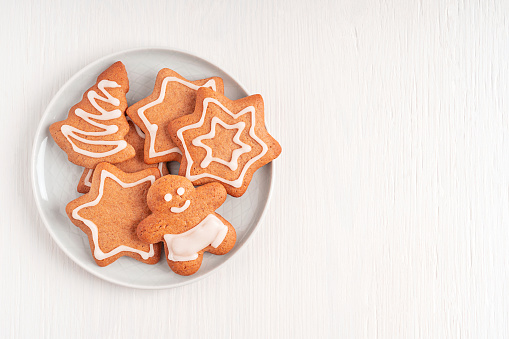Top view of homemade baked spiced gingerbread cookies or crunchy biscuits made with ginger spice and cinnamon decorated with sugar icing served on plate on white wooden background with copy space