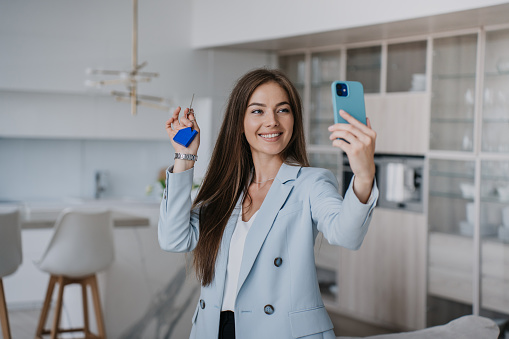 Pretty beautiful young woman in blue jacket shows keys of a new house to parents during video call by phone. Excited blonde businesswoman screams in happiness, purchased a flat. Real Estate Agent holds keys.