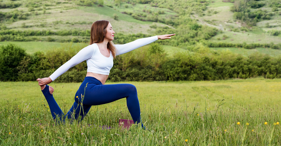 Young woman in sporty outfit exercising and stretching on mat in meadow outdoors. Hills landscape on background. Healthy active lifestyle