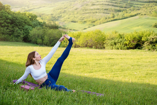 Young woman in sporty outfit exercising yoga and stretching on mat in meadow outdoors. Hills landscape on background. Healthy active lifestyle in summer