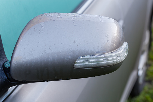 Side view mirror of the parked gray car with integrated turn signal repeater covered with dew, front view on a blurred background of misted side window and door close-up