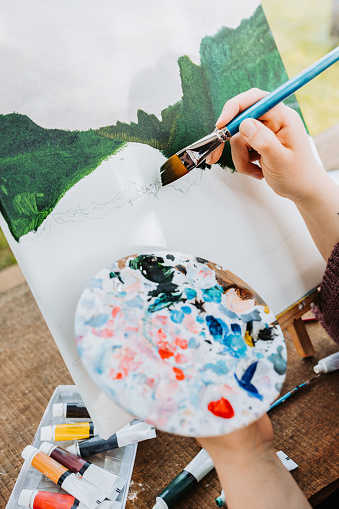 Unrecognizable young artist woman painting her own mountain scenery with oil on a canvas. Amateur painter.