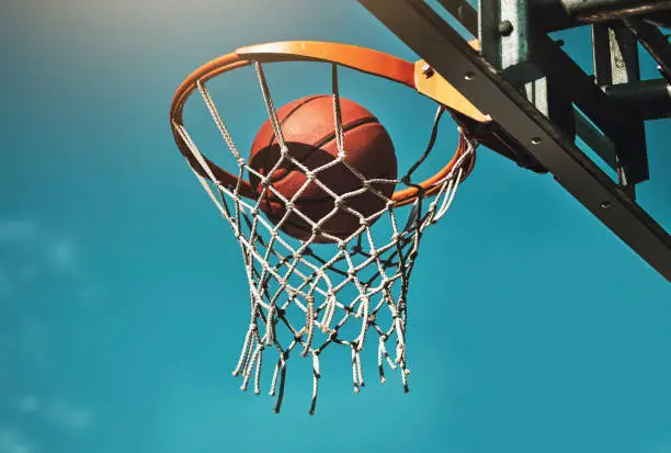 Basketball, goal and target in basket for sports match training on outdoor athletic court. Aim, score and winner with ball dunk in net at competition practice from low angle with blue sky.