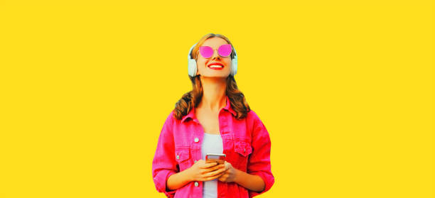 Portrait of stylish happy smiling young woman in headphones with smartphone listening to music wearing pink jacket, sunglasses on yellow background stock photo