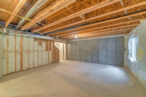 Unfinished basement with a plastic vapor barrier on the wall. Empty basement interior with wooden frameworks and insulated wall on the left and a window on the right.