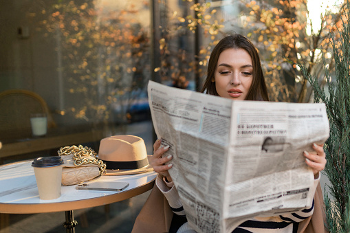 Beautiful girl in a coat reads a newspaper and smiles while relaxing in an outdoor cafe.