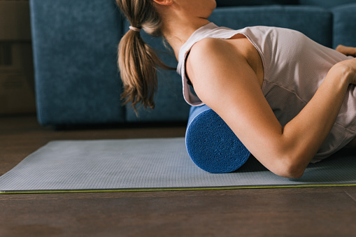 Caucasian brunette lying on a foam roller on a mat in her living room, stretching her back after a workout.