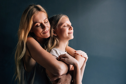 The love between a mother and daughter is forever: a smiling Caucasian mother embracing her daughter with eyes closed. (studio shot)