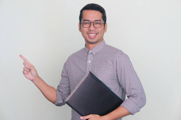 istock Adult Asian man smiling and pointing to the right side while holding laptop computer 1446790696