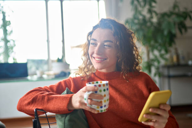 istock Young happy woman using mobile phone drinking coffee daydreaming at home. 1446806024