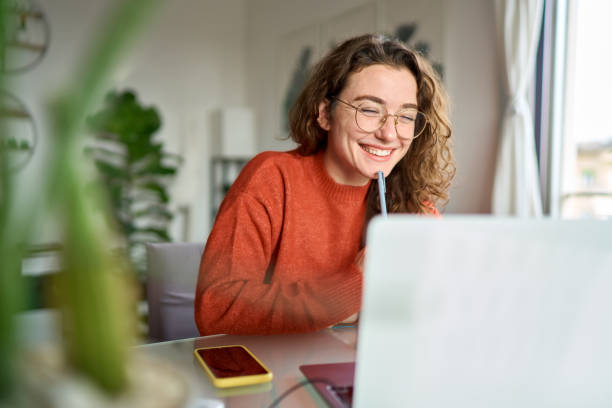 Young happy woman student using laptop watching webinar writing at home. Happy young woman using laptop sitting at desk writing notes while watching webinar, studying online, looking at pc screen learning web classes or having virtual call meeting remote working from home. looking over stock pictures, royalty-free photos & images