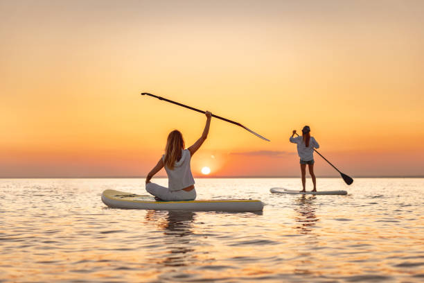 two happy girls walks at sunset lake on sup boards - paddle surfing stockfoto's en -beelden