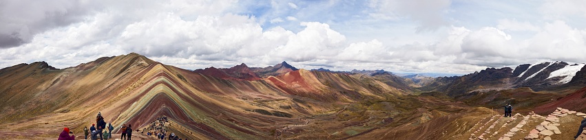 A panorama of the Rainbow mountains in Peru with people watching and could in the sky