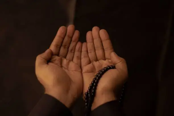A Muslim man raising his hands to pray with a Tasbeeh on a dark background