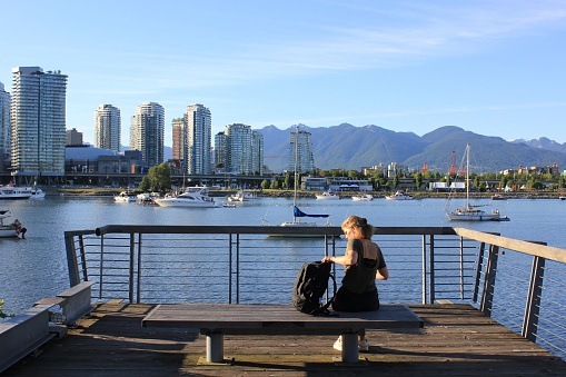 Vancouver, Canada – June 25, 2022: A young woman sitting on a bench at the waterfront of False Creek with boats