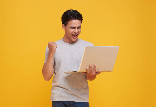 excited handsome young Asian man carrying laptop computer and raising his fist doing yes gesture isolated on yellow studio background. stock photo