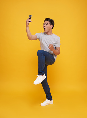 Happy young asian teen man celebrating with mobile phone isolated over yellow background.