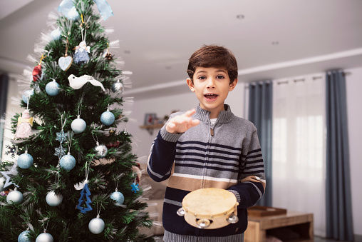 boy plays the tambourine next to his christmas tree