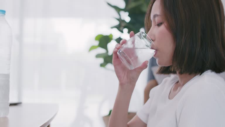 Young beautiful Asian woman drinking water from a drinking glass while sitting in the living room at home.