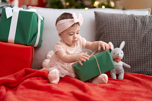 Adorable toddler opening christmas gift sitting on sofa at home