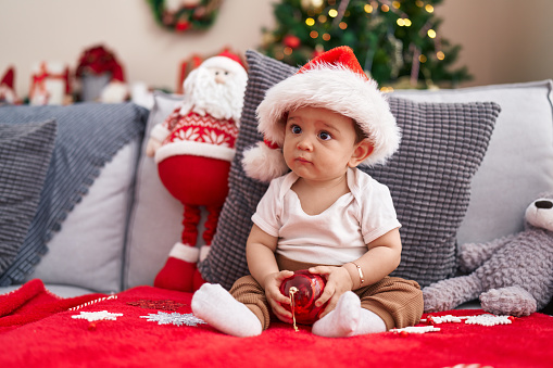 Adorable hispanic baby holding christmas decoration ball sitting on sofa at home