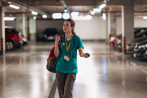 A young Caucasian female nurse is standing in a parking lot with a confused look on her face, holding out her car keys while searching for her car.