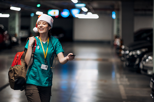 A young Caucasian female nurse wearing a Santa hat is cheerfully walking through an underground garage while searching for her car.