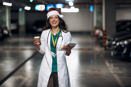 A young Caucasian female doctor wearing a Santa hat is looking at the camera, while cheerfully walking through an underground garage.