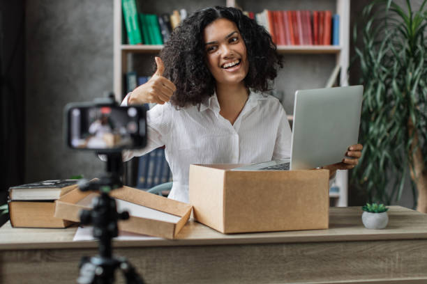 istock Multicultural woman recording video on phone camera while unpacking box with new wireless laptop. 1446878576