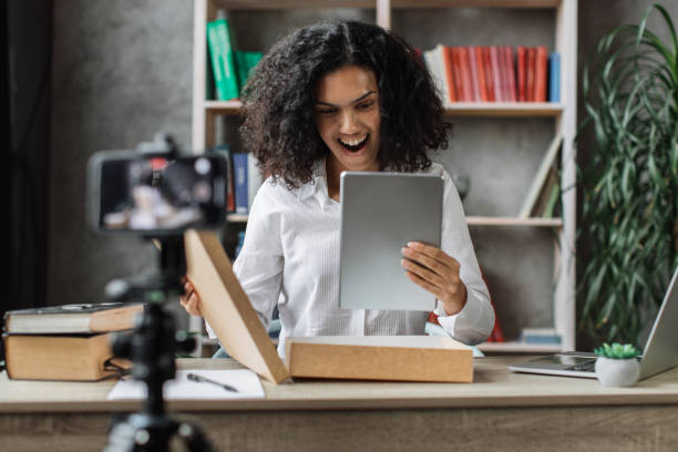 istock Multicultural woman recording video on phone camera while unpacking box with new wireless tablet. 1446878583